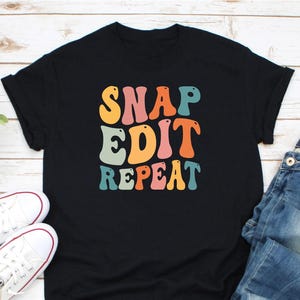 May include: Black t-shirt with the words "SNAP EDIT REPEAT" in colorful, retro-style lettering. The shirt is paired with white sneakers and blue jeans, creating a casual, everyday look.