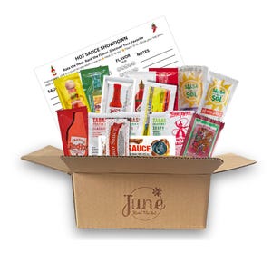May include: An open cardboard box filled with various hot sauce packets and small bottles. A sheet of paper with the text "HOT SAUCE SHOWDOWN" is visible. The box has the text "June Street Filadel" printed on it.