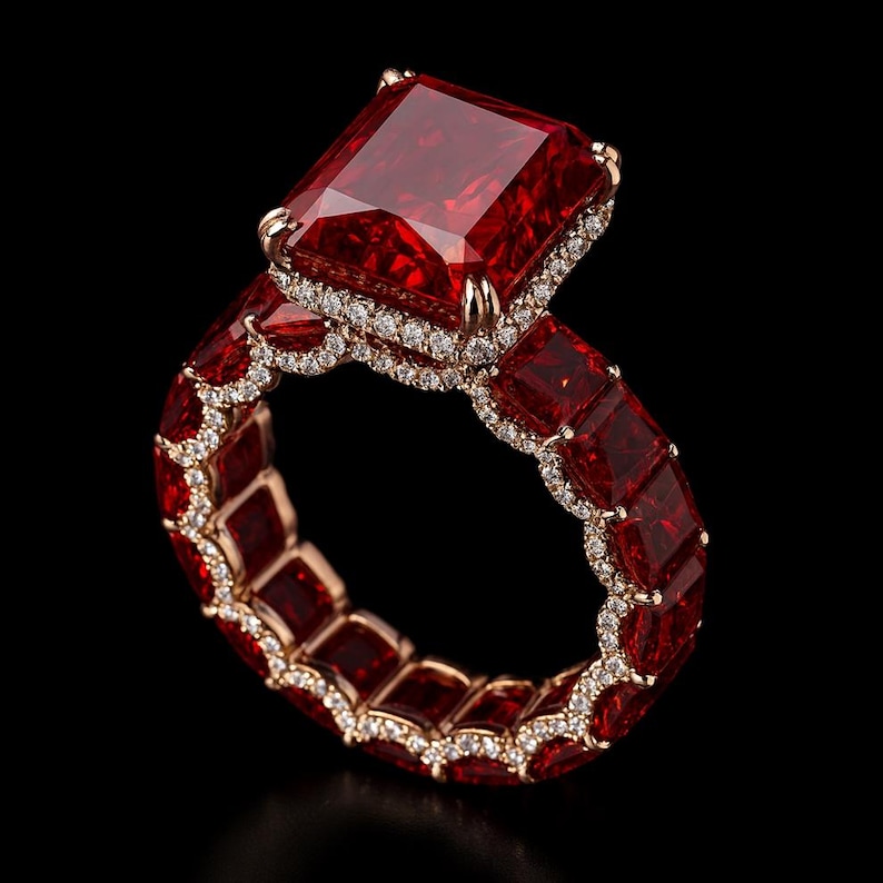 May include: A luxurious ring featuring a large, rectangular, deep red gemstone as the centerpiece. The ring's band is adorned with smaller, square-cut red stones and accented with a delicate border of small, clear gemstones. The setting is a warm gold color, creating a striking contrast.