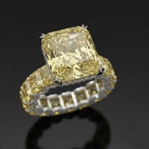 Puede incluir: Un lujoso anillo con un gran diamante amarillo de talla cuadrada. La banda del anillo está incrustada con diamantes amarillos más pequeños, creando un brillo continuo. El diseño se presenta sobre un fondo oscuro, resaltando el brillo del anillo.