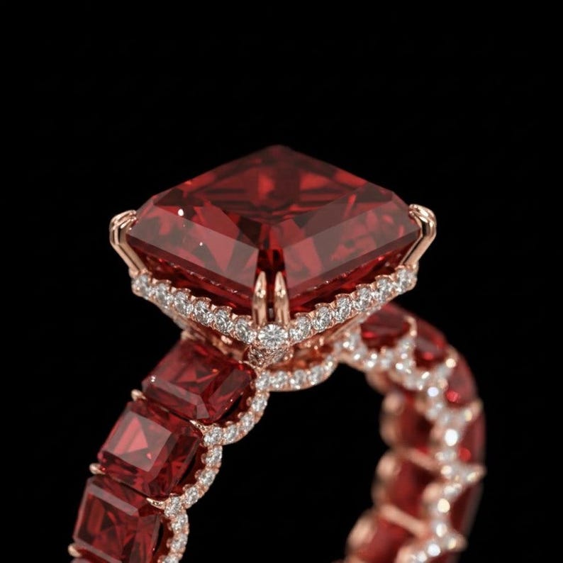 May include: A rose gold ring featuring a large, square-cut, deep red gemstone as the centerpiece. The ring is adorned with smaller red gemstones and a halo of small, clear stones around the main stone, set against a black background.
