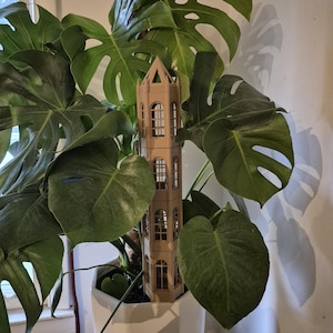May include: A tan, tower-shaped plant support structure with arched windows is nestled among the large, green leaves of a Monstera plant. The plant is in a white pot, and the scene is lit by natural light.