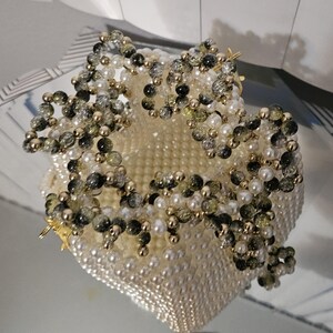 May include: A handcrafted, off-white beaded bag with a floral design. The bag is adorned with white pearls, gold beads, and clusters of green and black beads. The bag's interior is visible, and the overall design is elegant.