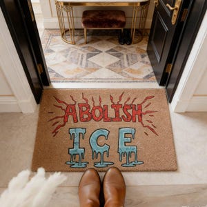Puede incluir: Una alfombra rectangular con las palabras "ABOLISH ICE" en rojo y azul. La alfombra está hecha de un material de fibra natural y se coloca frente a una puerta. Se ven zapatos marrones en la parte inferior de la imagen.