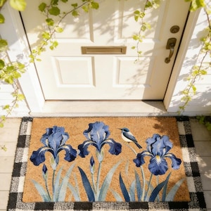 Op de afbeelding: Kokosmat met drie blauwe irissen en een kleine blauw-witte vogel. De mat heeft een natuurlijke bruine achtergrond en een zwart-wit geblokte rand. Een decoratieve welkomstmat voor een voordeur.