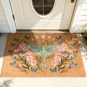 May include: A rectangular doormat with a nature-inspired design. The mat features a large, pale green moth in the center, surrounded by pink flowers, blue butterflies, and crescent moons. The mat is made of a natural brown material.