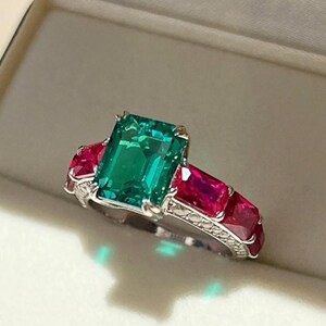 Può includere: Un anello color argento con una grande pietra preziosa verde taglio smeraldo al centro, affiancata da due pietre rosse taglio rettangolare su ciascun lato. La fascia in metallo presenta una superficie testurizzata lungo i lati.