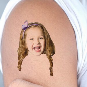 May include: A digitally created image of a smiling child with long, curly brown hair and a purple headband with a butterfly. The child's face is superimposed on a person's arm, with a white shirt visible in the background.