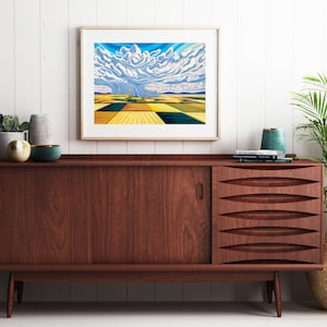 May include: A framed print of a landscape painting depicting a stormy sky and a field of crops. The painting is hanging on a white wall above a wooden sideboard.