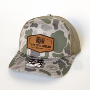 Może przedstawiać: Czapka typu trucker w kamuflażu z brązową skórzaną naszywką z napisem "TAYLOR FARMS EST. 1934" i grafiką traktora. Czapka ma siatkowy tył i zakrzywiony daszek. Czapka jest regulowana.