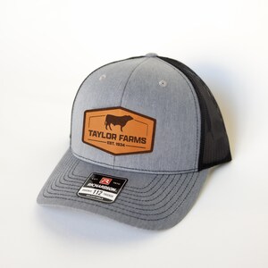 Może przedstawiać: Szara i czarna czapka typu trucker z brązową skórzaną naszywką z grafiką byka i napisem "TAYLOR FARMS EST. 1934". Czapka ma zakrzywiony daszek i regulowany pasek.
