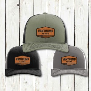 Może przedstawiać: Trzy czapki typu trucker z logo "Vantrump Farms". Czapki są w kolorze oliwkowym, czarnym i szarym, każda z siatkowym tyłem i brązową prostokątną naszywką. Logo zawiera tekst "Vantrump Farms" i wzór w paski.