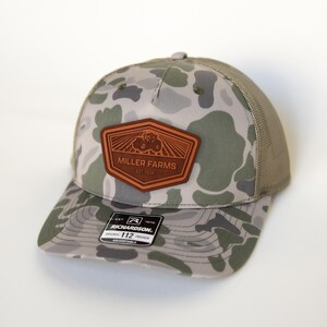 Może przedstawiać: Czapka typu trucker w kamuflażu z brązową skórzaną naszywką z napisem "MILLER FARMS EST 1934" i grafiką traktora. Czapka ma siatkowy tył i zakrzywiony daszek. Czapka typu trucker Richardson 112 jest regulowana.