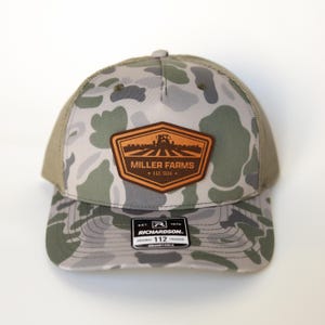 Może przedstawiać: Czapka typu trucker w kamuflażu z brązową naszywką z napisem "MILLER FARMS EST. 1934." Czapka ma siatkowy tył i zakrzywiony daszek. Na metce widnieje napis "RICHARDSON 112 ORIGINAL TRUCKER ADJUSTABLE."
