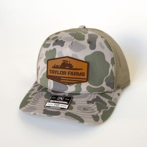 Może przedstawiać: Czapka typu trucker w kamuflażu z brązową skórzaną naszywką z napisem "TAYLOR FARMS" i grafiką traktora. Czapka ma siatkowy tył i zakrzywiony daszek. Etykieta głosi "RICHARDSON 112 TRUCKER ADJUSTABLE".