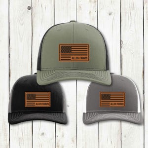 Puede incluir: Tres gorras de camionero en verde oliva, negro y gris. Cada gorra presenta un parche marrón con la bandera estadounidense y el texto "ALLEN FARMS" debajo. Las gorras tienen una parte trasera de malla y una visera curva.