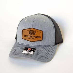 Może przedstawiać: Szara i czarna czapka typu trucker z brązową skórzaną naszywką. Na naszywce widnieje napis "TAYLOR FARMS" i grafika traktora, z napisem "EST. 1934" poniżej. Czapka ma zakrzywiony daszek i regulowany pasek.