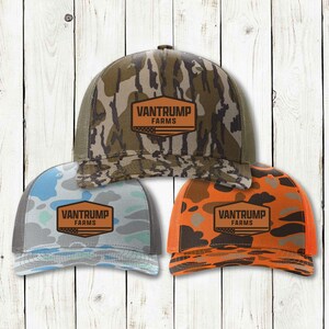 Puede incluir: Tres gorras de camionero con el texto "VANTRUMP FARMS" en un parche marrón. Una gorra tiene un patrón de camuflaje, otra tiene un patrón azul y gris, y la tercera tiene un patrón naranja y marrón. Todas tienen la parte trasera de malla.