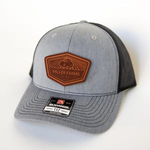 May include: Gray trucker hat with a black mesh back and a brown leather patch. The patch features a tractor graphic and the text "MILLER FARMS EST. 1934". The hat has a curved brim and an adjustable strap.
