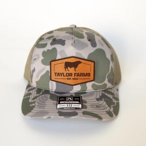 Może przedstawiać: Czapka typu trucker w kamuflażu z brązową skórzaną naszywką z sylwetką byka i napisem "TAYLOR FARMS EST. 1934". Czapka ma siatkowy tył i zakrzywiony daszek. Etykieta głosi "RICHARDSON 112 TRUCKER".