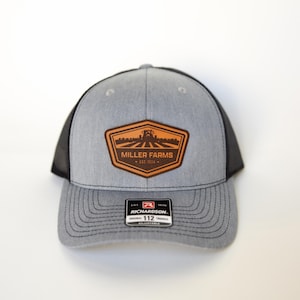 Puede incluir: Gorra de camionero gris y negra con una etiqueta Richardson. La gorra presenta un parche de cuero marrón con las palabras "MILLER FARMS EST. 1934" y una escena de granja. La gorra es ajustable.