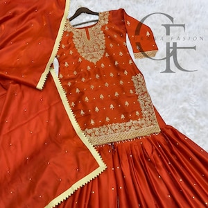 May include: A vibrant orange three-piece outfit consisting of a long-sleeved tunic, wide-leg pleated trousers, and a matching dupatta. The tunic features intricate gold embroidery around the neckline and hem. The text GURAWA FASION is visible.