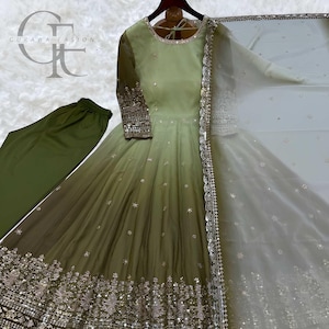 May include: An olive green ombre anarkali dress with long sleeves, featuring intricate silver embroidery and sequins along the hem and cuffs. The set includes matching olive green trousers and a sheer white dupatta with a silver embroidered border.