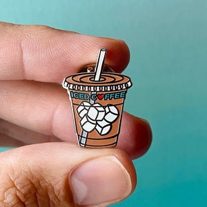 Puede incluir: Un pin de esmalte marrón y blanco de una taza de café helado con un corazón rojo y el texto "ICED COFFEE".