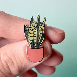 Puede incluir: Un pin de esmalte dorado con una planta serpiente en maceta con hojas verdes y amarillas. La planta está en una maceta de color terracota.
