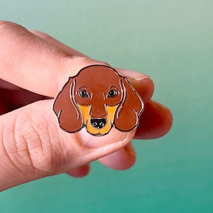 May include: A brown and tan dachshund enamel pin. The pin features a close-up of the dog's face.