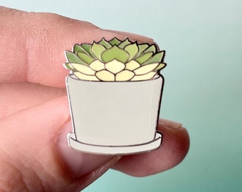 May include: A white enamel pin depicting a green succulent plant in a white pot. The pin is a charming and whimsical representation of a houseplant.