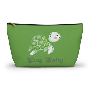May include: A green zippered pouch featuring a cartoon turtle with a green shell and white accents. The text "Stay Salty" is printed in gray below the turtle. The pouch has a black zipper and a gold-colored pull.