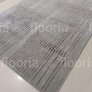 May include: A rectangular gray rug with a linear pattern. The rug features a series of parallel lines running across its surface, creating a textured effect. The rug is made of a soft material and is suitable for living rooms and bedrooms.