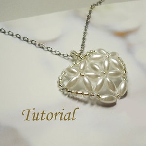 Pode incluir: Um colar de corrente prateada com um pingente de coração de pérolas brancas. O pingente é feito de pequenas pérolas brancas e contas prateadas dispostas em um padrão de flores. A palavra "Tutorial" está escrita em letras douradas abaixo do pingente.