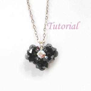 May include: A beaded necklace with a heart-shaped pendant. The pendant is made of black beads and features a central iridescent crystal. The necklace has a silver chain. The word "Tutorial" is written in pink.