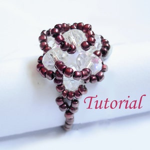 Pode incluir: Um close-up de um anel de contas com um design de cor bordô e cristal transparente. O anel está sobre uma superfície branca com a palavra "Tutorial" escrita em rosa.