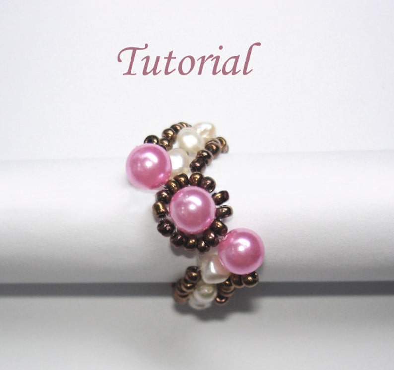 Op de afbeelding: Een roze en witte kralenring met bruine zaadkraaltjes. De ring is gemaakt met een geweven patroon en heeft verschillende ronde roze kralen.