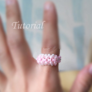 May include: A pink and white beaded ring worn on a finger. The ring is made of small, round beads that are strung together in a spiral pattern. The beads are alternating pink and white colors.
