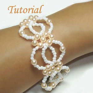 May include: A handcrafted bracelet with a design of white beaded circles and peach-coloured pearl beads. The word "Tutorial" is written in orange script at the top. The bracelet is worn on a wrist.