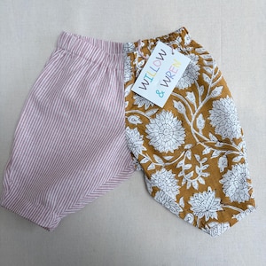 May include: Baby bloomers with a split design. One side has vertical pink and white stripes, the other a mustard yellow floral pattern with white flowers. A tag reads "WILLOW & WREN".