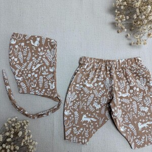 May include: A brown baby bonnet and shorts set with a white woodland animal and floral pattern. The set includes a bonnet with ties and elastic waist shorts. The fabric has a soft, textured appearance.