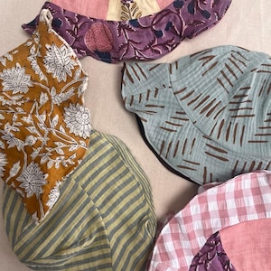May include: A selection of patterned sun hats in various colours. The hats feature floral prints, stripes, and gingham patterns. Colours include pink, yellow, blue, and purple. The hats are made of fabric and appear to be handmade.