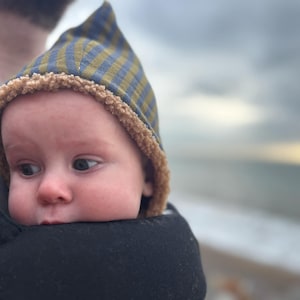 May include: A baby wearing a blue and gold check patterned hat with a brown sherpa trim, held in a black baby carrier. The baby gazes to the side. The background shows a blurred view of the sea and sky.