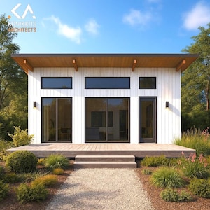Op de afbeelding: Moderne witte cabine met een houten dek en een grindpad dat naar de ingang leidt. De cabine heeft grote ramen, een plat dak en de tekst "KUNIKKIS ARCHITECTS". De landschapsarchitectuur omvat struiken en bloemen.