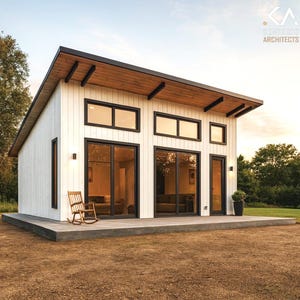 Puede incluir: Cabaña blanca moderna con marcos negros y un tejado inclinado. Grandes ventanas y puertas correderas de cristal proporcionan abundante luz natural. Una mecedora de madera se encuentra en el patio de hormigón. La cabaña está rodeada de árboles y césped.
