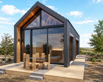 May include: Modern cabin with a dark gray exterior, wooden accents, and large glass windows. A wooden deck with a table and chairs extends from the cabin. The cabin is set against a backdrop of a blue sky and green landscape.