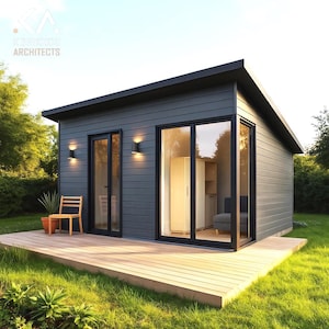 May include: A modern, gray-sided shed with a black roof and trim. Large glass doors and windows reflect the interior. A wooden deck extends from the building, with a chair and potted plant visible. The text "KOKIKKIE ARCHITECTS" is in the upper left corner.