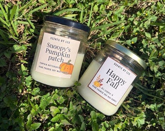 May include: Two clear glass jars filled with off-white soy wax candles. The left label reads Scents by Lex, Snoopy's Pumpkin Patch, Pumpkin Buttercream, Hand Poured Soy Wax Candle. The right label reads Scents by Lex, Happy Fall, All Leaves 'R, Hand Poured Soy Wax Candle.