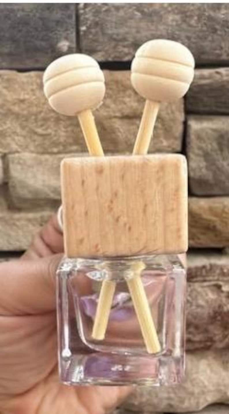 May include: A clear, square glass bottle with a wooden top and two wooden diffuser sticks. The top has two spherical wooden accents. The bottle is held in a hand.