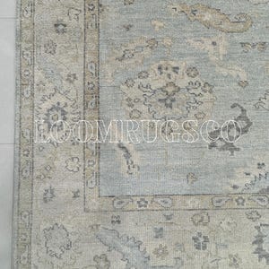 Pode incluir: Uma secção de um tapete com padrão azul claro e bege com uma borda detalhada. O tapete apresenta um design central com motivos florais e geométricos. O texto "LOOMIRUGSCO" é visível na parte inferior do tapete.